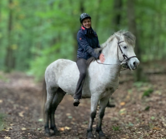 Eine Reiterin auf einem weißen Pferd in einem grünen Wald, beide entspannt und ruhig. 